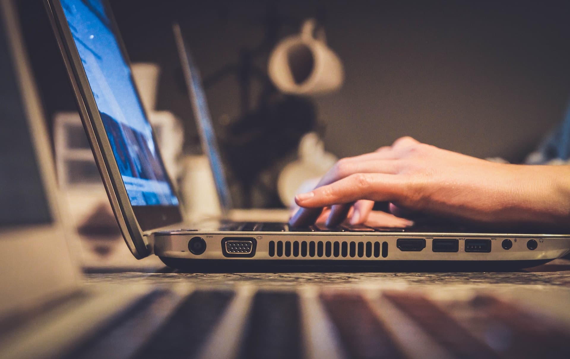 A imagem mostra um close-up da lateral de um laptop prateado em uma mesa. O foco principal está na mão de uma pessoa digitando no teclado. Apenas parte da mão está visível, com destaque para os dedos sobre as teclas. A tela do computador está ligada e exibe conteúdo, mas está fora de foco e não é possível distinguir o que está sendo mostrado. Ao fundo, há objetos borrados que sugerem um ambiente de escritório ou uma casa, mas não são claros o suficiente para identificação detalhada. A iluminação é suave e quente, criando uma atmosfera confortável e concentrada.